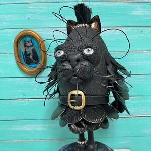 May include: A quirky black cat sculpture with blue eyes, whiskers, and a belt with a gold buckle. The cat is mounted on a wooden base and has a small framed painting of a cat in the background. The sculpture has a unique, artistic design.