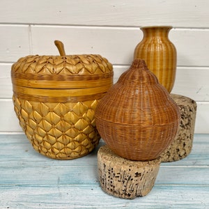 May include: Three woven baskets in different shapes and sizes. The largest basket is shaped like an apple and has a lid. The middle basket is shaped like a pear. The smallest basket is a cylindrical vase.