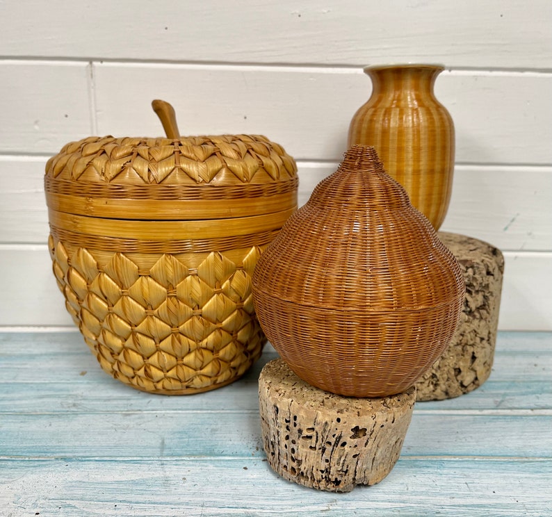 May include: Three woven baskets in different shapes and sizes. The largest basket is shaped like an apple and has a lid. The middle basket is shaped like a pear. The smallest basket is a cylindrical vase.