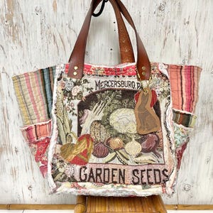 May include: A large tote bag with brown leather handles and a decorative fabric design. The fabric features a vintage-style illustration of vegetables and the words "GARDEN SEEDS" and "MERCERSBURG PA". A leather tag with a rabbit silhouette hangs from the handle.