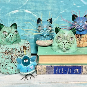 May include: Four whimsical ceramic cat figurines with colorful wire whiskers, sitting on a stack of books. The cats are painted in shades of blue and green, with expressive faces and detailed features. The books are covered in a brown paper with a title label that reads "Two Years before the Mast."