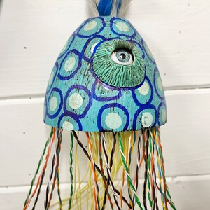 May include: A blue and white painted wooden jellyfish with a large blue eye and a long, colorful string of tentacles.