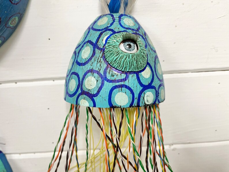 May include: A blue and white painted wooden jellyfish with a large blue eye and a long, colorful string of tentacles.