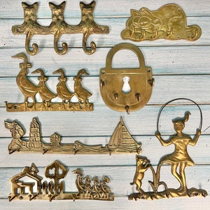 Seven different brass key racks done in a flatlay style. There are cats, Garfield, 4 ducks, a padlock, a lighthouse with sailboat, a little house with ducks in the yard and a girl skipping rope with her dog. Housewarming gift at CastawaysHall on Etsy