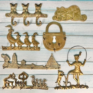 Seven different brass key racks done in a flatlay style. There are cats, Garfield, 4 ducks, a padlock, a lighthouse with sailboat, a little house with ducks in the yard and a girl skipping rope with her dog. Housewarming gift at CastawaysHall on Etsy