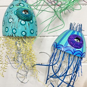 Two jellyfish sculptures by CastawaysHall hang on a wall. They are painted in blues and turquoise and are made out of wood net floats. They have brightly coloured wire tentacles and one blue eye surrounded by sculpted clay.