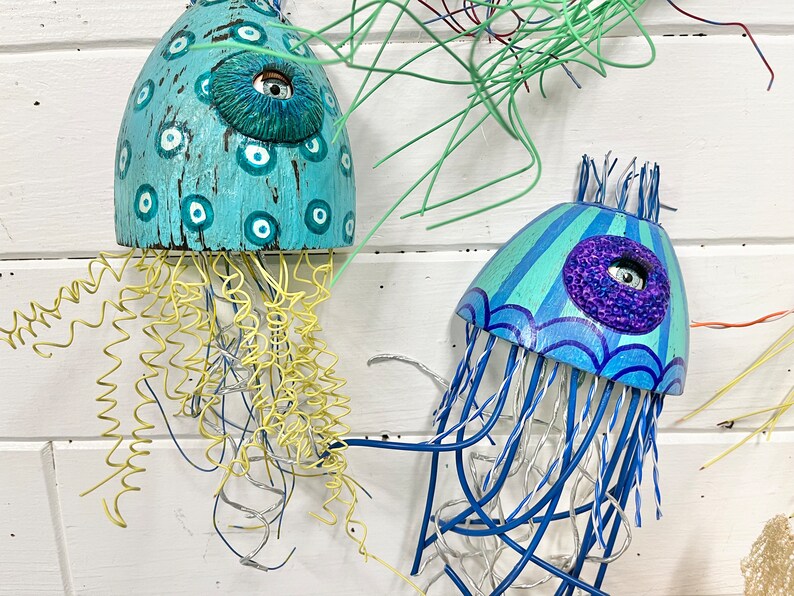 Two jellyfish sculptures by CastawaysHall hang on a wall. They are painted in blues and turquoise and are made out of wood net floats. They have brightly coloured wire tentacles and one blue eye surrounded by sculpted clay.