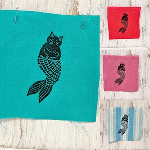 Peut inclure: Un carré de tissu turquoise avec un motif de chat-sirène noir, épinglé sur une surface en bois blanc. Des carrés de tissu supplémentaires en rouge, rose et bleu rayé présentent le même motif. Le chat-sirène a une tête de chat et une queue de poisson.