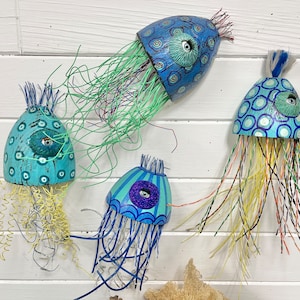Four jellyfish sculptures by CastawaysHall hang on a wall. They are painted in blues and turquoise and are made out of wood net floats. They have brightly coloured wire tentacles and one blue eye surrounded by sculpted clay.