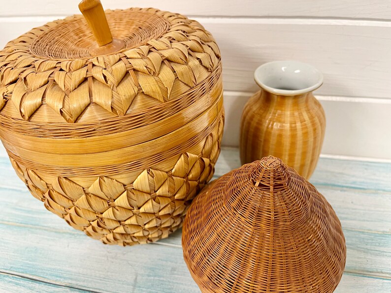 May include: Three woven baskets made of natural materials. The largest basket is shaped like an apple and has a lid. The middle basket is a tall vase with a brown and white striped pattern. The smallest basket is a cone shape.