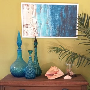 K&ouml;nnte beinhalten: Ein Holzrahmenbild mit einer abstrakten Ozeanszene in Blau-, Gr&uuml;n- und Wei&szlig;t&ouml;nen. Drei blaue Glasflaschen mit unterschiedlichen Formen und Texturen sind auf einem Holztisch mit einer gro&szlig;en Muschel und einer Glasglocke angeordnet.