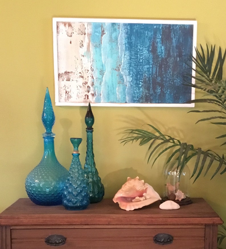K&ouml;nnte beinhalten: Ein Holzrahmenbild mit einer abstrakten Ozeanszene in Blau-, Gr&uuml;n- und Wei&szlig;t&ouml;nen. Drei blaue Glasflaschen mit unterschiedlichen Formen und Texturen sind auf einem Holztisch mit einer gro&szlig;en Muschel und einer Glasglocke angeordnet.