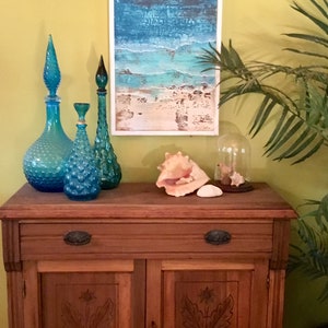 K&ouml;nnte beinhalten: Ein Holzschr&auml;nkchen mit zwei T&uuml;ren und einer Schublade. Der Schrank hat geschnitzte Blumendekore auf den T&uuml;ren. Auf dem Schrank stehen drei blaue Glasflaschen und eine Muschel. An der Wand hinter dem Schrank h&auml;ngt ein gerahmtes Bild mit einer Strandlandschaft.