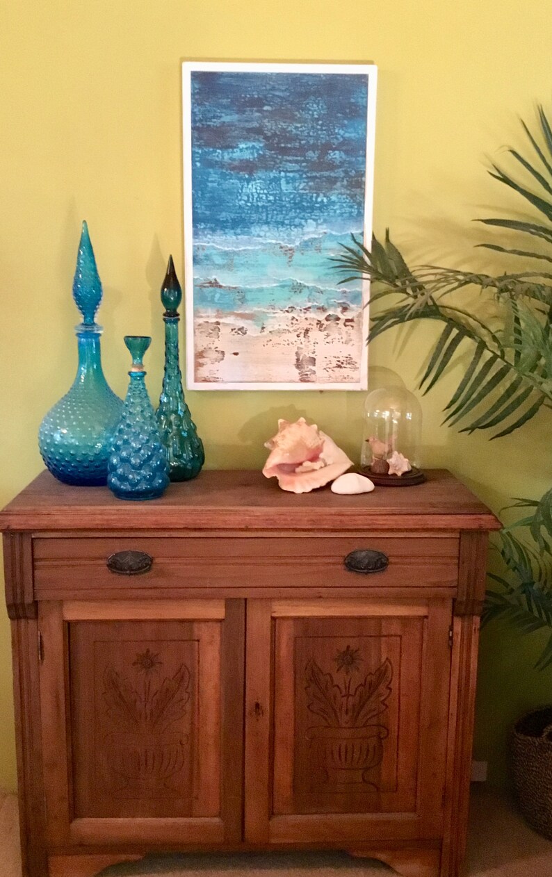 K&ouml;nnte beinhalten: Ein Holzschr&auml;nkchen mit zwei T&uuml;ren und einer Schublade. Der Schrank hat geschnitzte Blumendekore auf den T&uuml;ren. Auf dem Schrank stehen drei blaue Glasflaschen und eine Muschel. An der Wand hinter dem Schrank h&auml;ngt ein gerahmtes Bild mit einer Strandlandschaft.