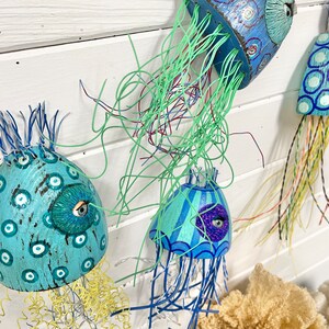 Four jellyfish sculptures by CastawaysHall hang on a wall. They are painted in blues and turquoise and are made out of wood net floats. They have brightly coloured wire tentacles and one blue eye surrounded by sculpted clay.