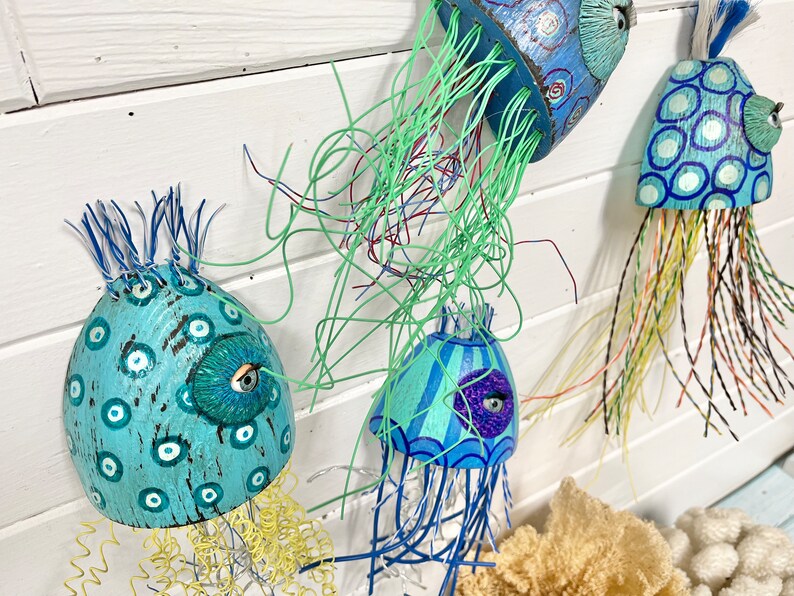 Four jellyfish sculptures by CastawaysHall hang on a wall. They are painted in blues and turquoise and are made out of wood net floats. They have brightly coloured wire tentacles and one blue eye surrounded by sculpted clay.