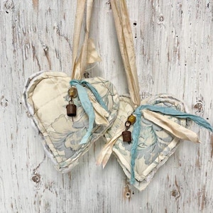 May include: Two heart-shaped fabric ornaments with a floral pattern in cream and blue. Each heart has a cream-colored trim, a blue ribbon, and a small bell. The ornaments hang from cream-colored ribbons against a white wooden background.