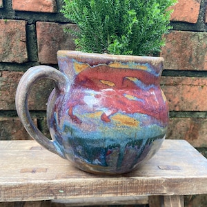 May include: A handmade ceramic planter with a brown, red, blue, and green glaze. The planter has a rounded body and a handle. A small green plant is growing in the planter.