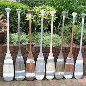 Peut inclure: Neuf pagaies en bois peintes dans différentes nuances de gris, blanc et marron. Les pagaies sont disposées en rangée sur une surface en briques.