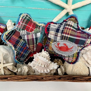 May include: A wicker basket containing decorative items. Two star-shaped cushions with plaid fabric and blue trim are visible, one with a crab patch. The basket also holds seashells, a starfish, and a plush animal.