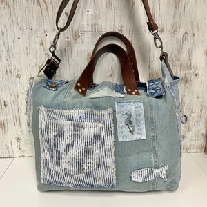 May include: A denim tote bag with brown leather handles and a detachable shoulder strap. The bag features a large front pocket with a white and blue striped design, and patches with seahorse and fish designs.