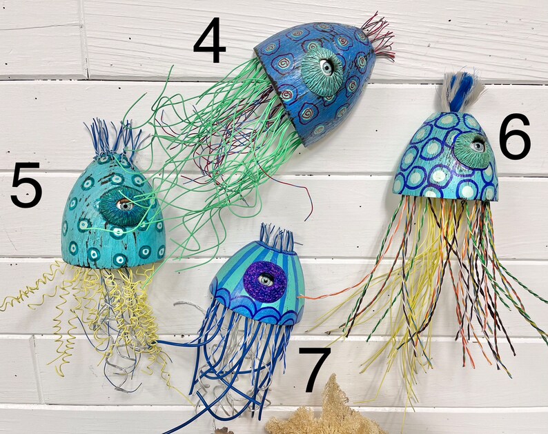 Four jellyfish sculptures by CastawaysHall hang on a wall. They are painted in blues and turquoise and are made out of wood net floats. They have brightly coloured wire tentacles and one blue eye surrounded by sculpted clay.