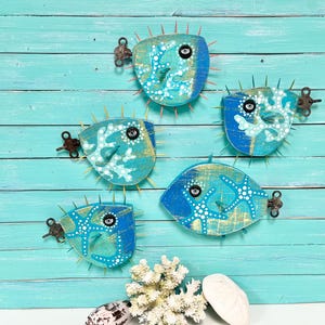 May include: Five wooden pufferfish ornaments painted in blue, white and gold. Each fish has a unique design and features a small metal keyhole. The ornaments are displayed against a turquoise blue wooden background.