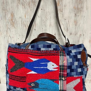 May include: A large, patchwork tote bag with a vibrant design. The bag features a red panel with blue and white fish, denim, and plaid accents. It has brown leather handles and a shoulder strap, perfect for carrying essentials.