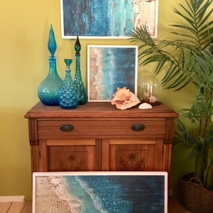 K&ouml;nnte beinhalten: Drei blaue Glasflaschen mit einer strukturierten Oberfl&auml;che und einem hohen, schlanken Hals, angeordnet auf einem Holzkasten mit drei gerahmten Bildern zum Thema Meer dar&uuml;ber. Die Bilder zeigen blaue und wei&szlig;e Farbt&ouml;ne mit einer strukturierten Oberfl&auml;che. Eine kleine, klare Glaskuppel steht auf dem Kasten mit einer Muschel darin.
