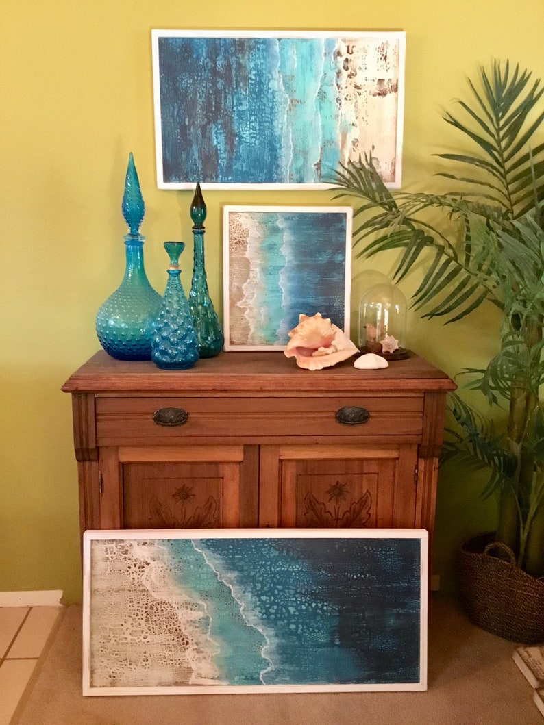 K&ouml;nnte beinhalten: Drei blaue Glasflaschen mit einer strukturierten Oberfl&auml;che und einem hohen, schlanken Hals, angeordnet auf einem Holzkasten mit drei gerahmten Bildern zum Thema Meer dar&uuml;ber. Die Bilder zeigen blaue und wei&szlig;e Farbt&ouml;ne mit einer strukturierten Oberfl&auml;che. Eine kleine, klare Glaskuppel steht auf dem Kasten mit einer Muschel darin.