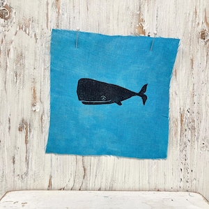 May include: A square of turquoise fabric with a black whale illustration pinned to a distressed white wooden surface. The whale is depicted in a simple, stylized form, with a rounded body and a small tail. The fabric has a raw edge.