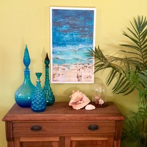 K&ouml;nnte beinhalten: Ein Holzschr&auml;nkchen mit zwei T&uuml;ren und zwei Schubladen, dekoriert mit blauen Glasflaschen und Muscheln. Ein Gem&auml;lde mit einer Strandszene h&auml;ngt &uuml;ber dem Schr&auml;nkchen.