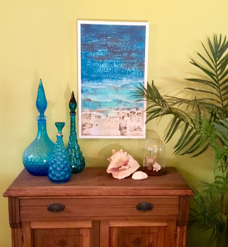 K&ouml;nnte beinhalten: Ein Holzschr&auml;nkchen mit zwei T&uuml;ren und zwei Schubladen, dekoriert mit blauen Glasflaschen und Muscheln. Ein Gem&auml;lde mit einer Strandszene h&auml;ngt &uuml;ber dem Schr&auml;nkchen.
