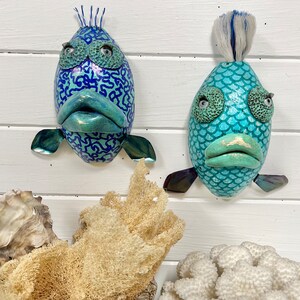 May include: Two whimsical fish sculptures with blue and green paint and textured surfaces. One fish has a patterned design and the other has a scaly design.