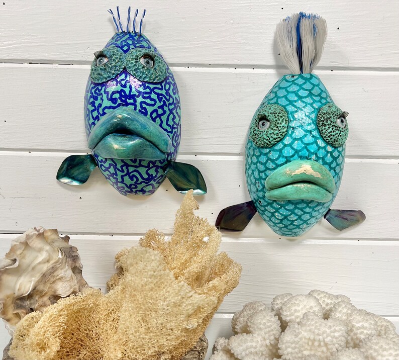 May include: Two whimsical fish sculptures with blue and green paint and textured surfaces. One fish has a patterned design and the other has a scaly design.