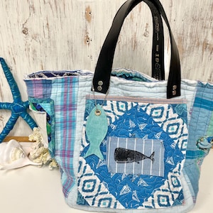 May include: A quilted tote bag featuring a patchwork of light blue, white, and purple plaid fabric. The front pocket displays a blue diamond pattern with a small rectangular patch showing a black whale illustration and a hanging teal fish charm.