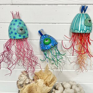 May include: Three handmade jellyfish wall decorations. The jellyfish are made of wood and painted in bright colors. One is blue and green with a single eye, another is blue and green with a single eye, and the third is teal and white with stripes and a single eye. The jellyfish are hanging from a white wall.