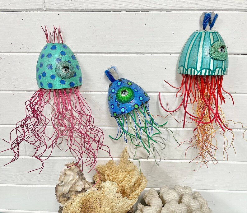 May include: Three handmade jellyfish wall decorations. The jellyfish are made of wood and painted in bright colors. One is blue and green with a single eye, another is blue and green with a single eye, and the third is teal and white with stripes and a single eye. The jellyfish are hanging from a white wall.