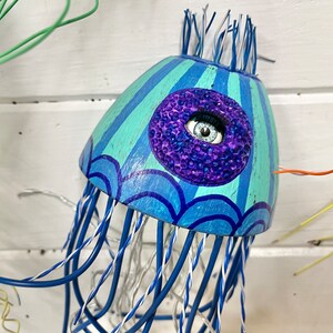 May include: A blue and green wooden jellyfish with a purple and blue eye. The jellyfish has a wire frame and is decorated with blue and white stripes.