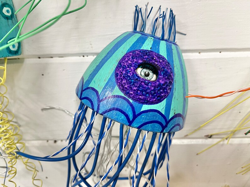 May include: A blue and green wooden jellyfish with a purple and blue eye. The jellyfish has a wire frame and is decorated with blue and white stripes.