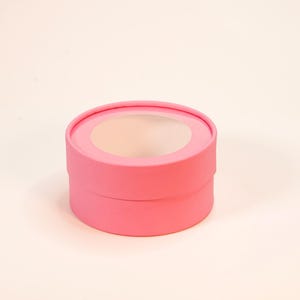 Könnte beinhalten: Eine runde, leuchtend rosa Geschenkbox aus Pappe mit einem kreisförmigen Sichtfenster im Deckel. Der Behälter hat eine glatte, matte Oberfläche und eine zylindrische Form mit einem abnehmbaren Deckel, der den Innenraum freigibt.
