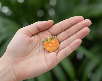 May include: A round orange enamel pin featuring two green leaves at the top and the white text "MEET ME IN MONTAUK" printed in the center. The pin has a gold-toned metal outline and a smooth, glossy finish, showcasing a vibrant citrus fruit design.