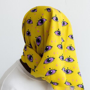 Puede incluir: Un pañuelo amarillo con un patrón repetido de ojos morados y formas de estrellas rosas. La tela está drapeada sobre la cabeza y los hombros de una persona, creando un efecto en capas. El fondo es blanco liso.