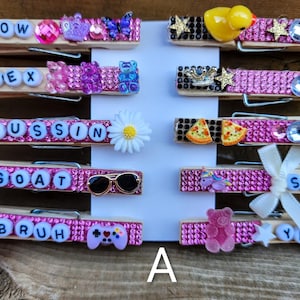 Puede incluir: Una colección de pinzas de ropa decorativas de madera, cada una adornada con purpurina rosa, cuentas y abalorios. Las pinzas presentan varias palabras deletreadas con cuentas de letras, incluyendo "GLOW", "FLEX" y "GOAT". Cada pinza está adornada con lazos y abalorios.