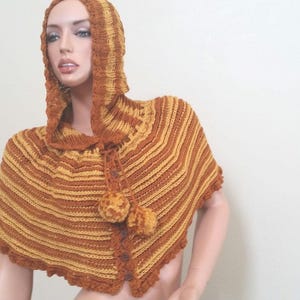 May include: A hooded knitted poncho with a striped pattern in shades of brown and yellow. The poncho has a crocheted trim and pom-poms.