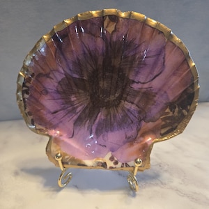 May include: A decorative scallop shell featuring a purple and black floral design on its interior surface. The edges of the shell are painted with a metallic gold finish. The shell is displayed upright on a small, gold-toned metal wire stand.