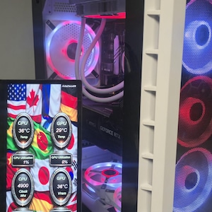 Puede incluir: Un ordenador personalizado con un panel lateral de cristal, que muestra los componentes internos y los ventiladores LED. Una pequeña pantalla muestra las métricas de rendimiento del sistema, incluyendo las temperaturas de la CPU y la GPU. La carcasa tiene un borde blanco.