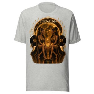May include: Light gray t-shirt featuring a gold illustration of two women, the Gemini zodiac sign, with musical notes and vinyl records. The word "GEMINI" is arched above the figures. The design has a celestial theme.