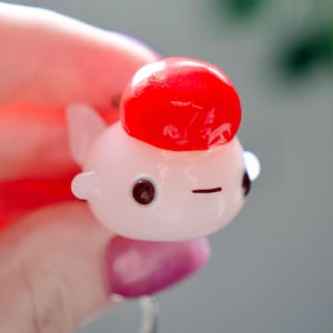 May include: A small, pink and red fish-shaped keychain. The fish has a red, rounded top, black eyes, and a small, horizontal black line for a mouth. The keychain is held by a silver ring.
