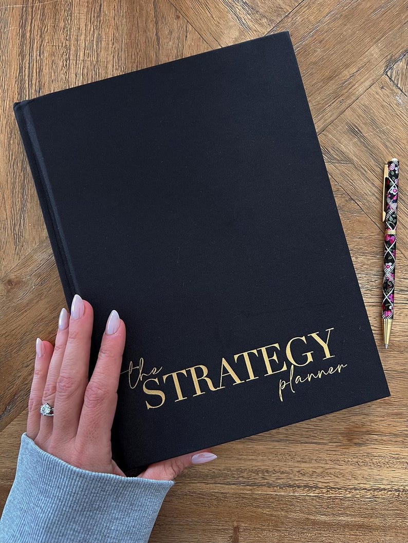 May include: A black Strategy Planner with gold lettering and a matching pen. The planner is held by a person. The pen has a floral design with black, pink, and gold accents. The planner is on a wooden surface.